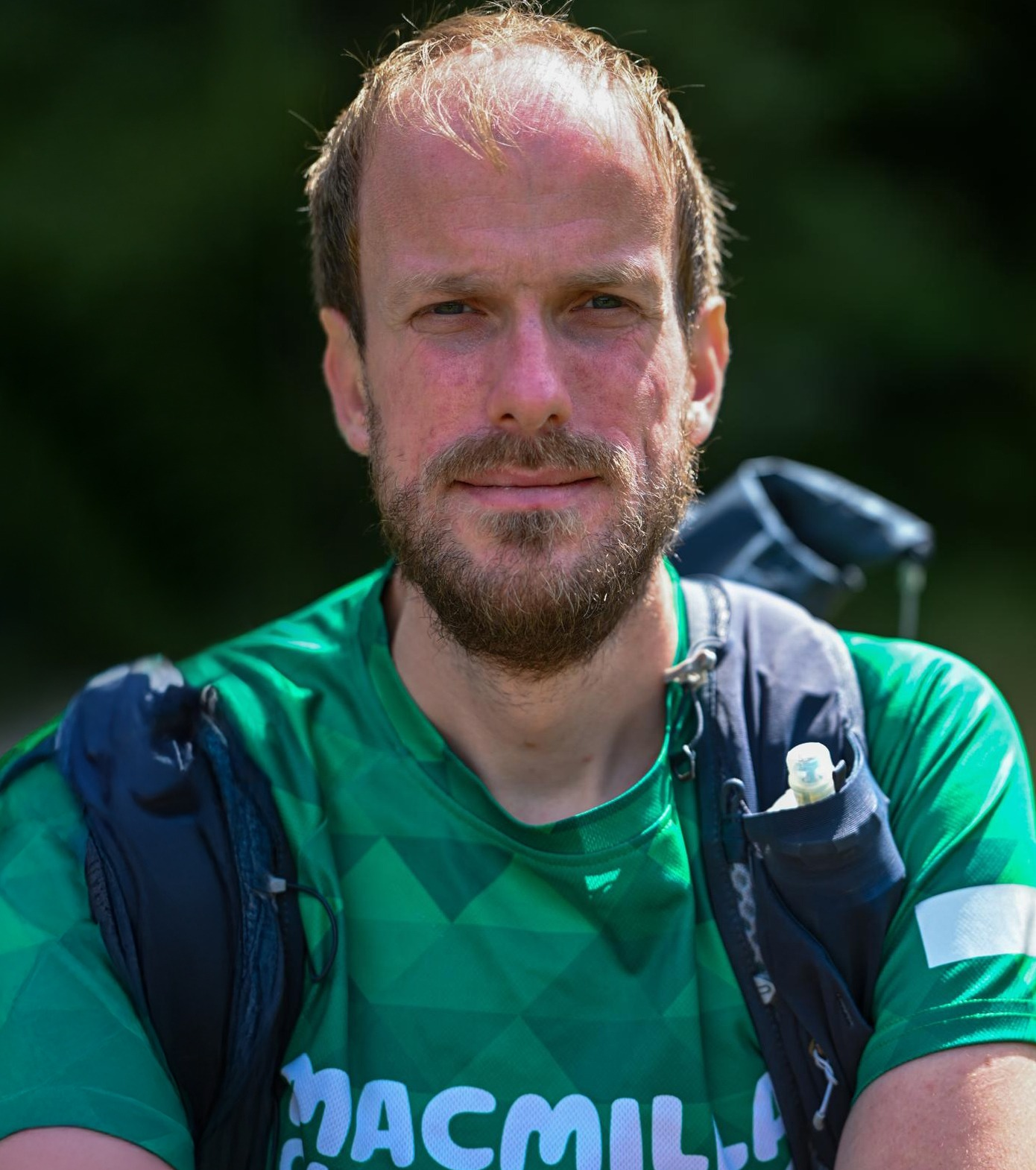 A photo of Nathaniel wearing a green Macmillan-branded athletic shirt stands outdoors with a hydration backpack strapped over his shoulders. The background is a natural, green, out-of-focus setting.