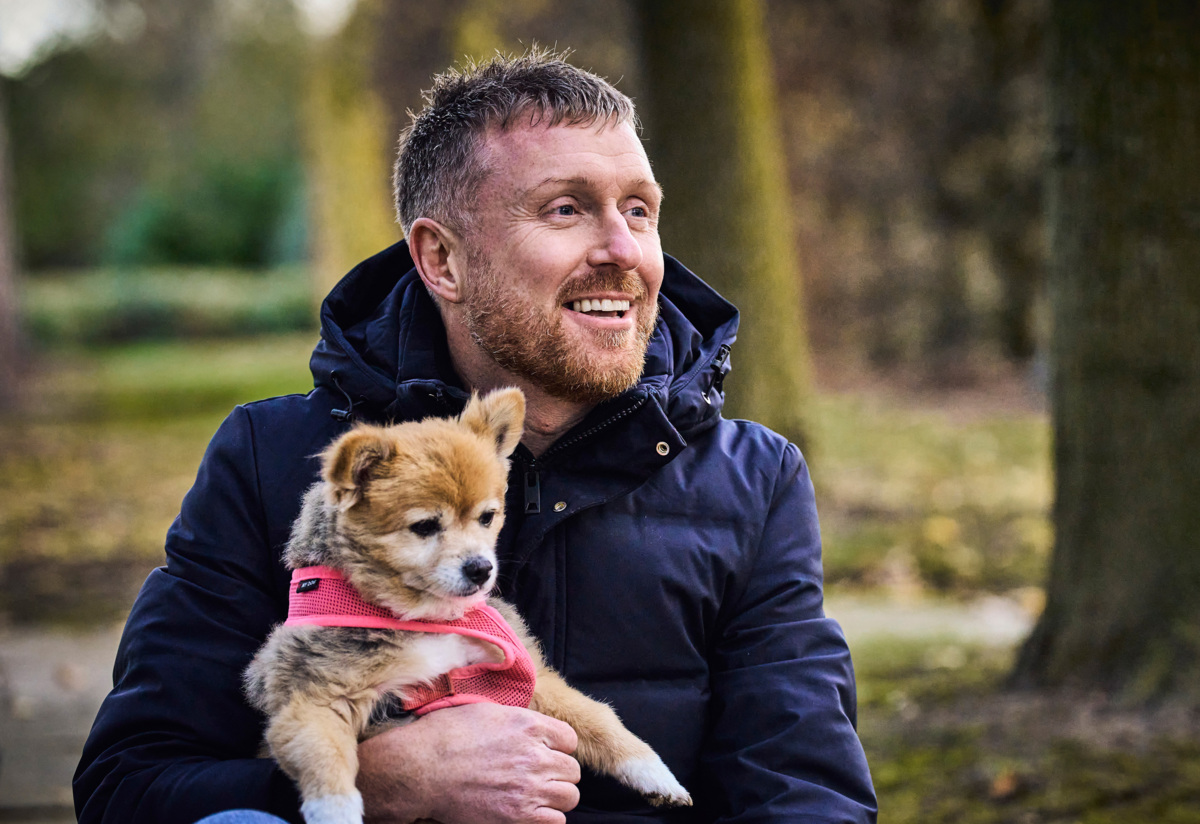 A smiling man sitting in a park with his small dog on his lap