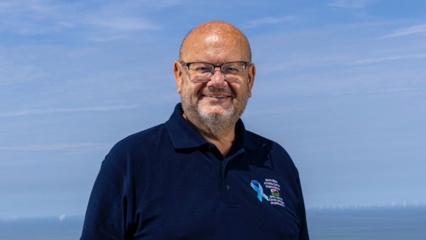 Neil is standing outside in the sunshine with the sea behind him. He is smiling and wearing glasses and a top with the North Wales Prostate Cancer Support Group name, a Welsh dragon, and a blue ribbon on it. 