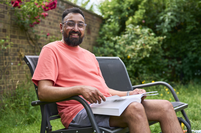 Mohammad sits on a garden bench with a book on his lap. He had a beard and wears glasses and is smiling.