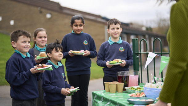 A group of school children are standing outside near a table decorated for a Coffee Morning. The children are all in a school uniform and are holding plates with food on them. 