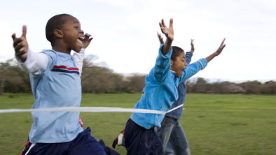 A group of children are running across a white finish line at an event. Their hands are in the air and they appear outside. Each of them are wearing blue tops and blue trousers.