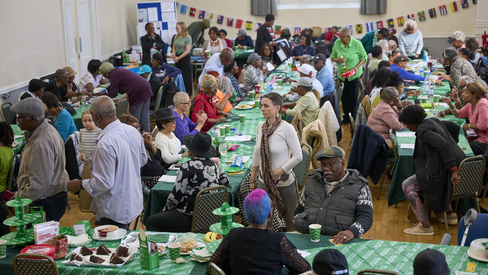 A large community centre is full of people for a Coffee Morning. There are table set up in rows for people to sit and enjoy food and drink. 