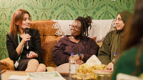 Three women are sitting on a brown sofa in conversation. One is holding a microphone and wearing a black dress. She is also wearing a green lanyard.