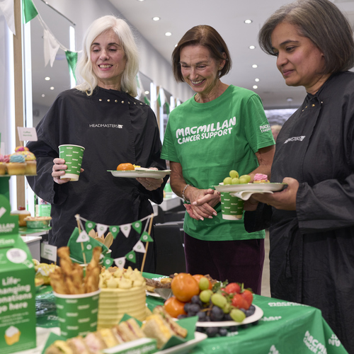 Three people are standing near a table that has been set up for a Coffee Morning. They are in a hair dressers. Two of the people are wearing hair cutting capes. The table has different food items on it like yummy fresh fruit and moreish cakes.