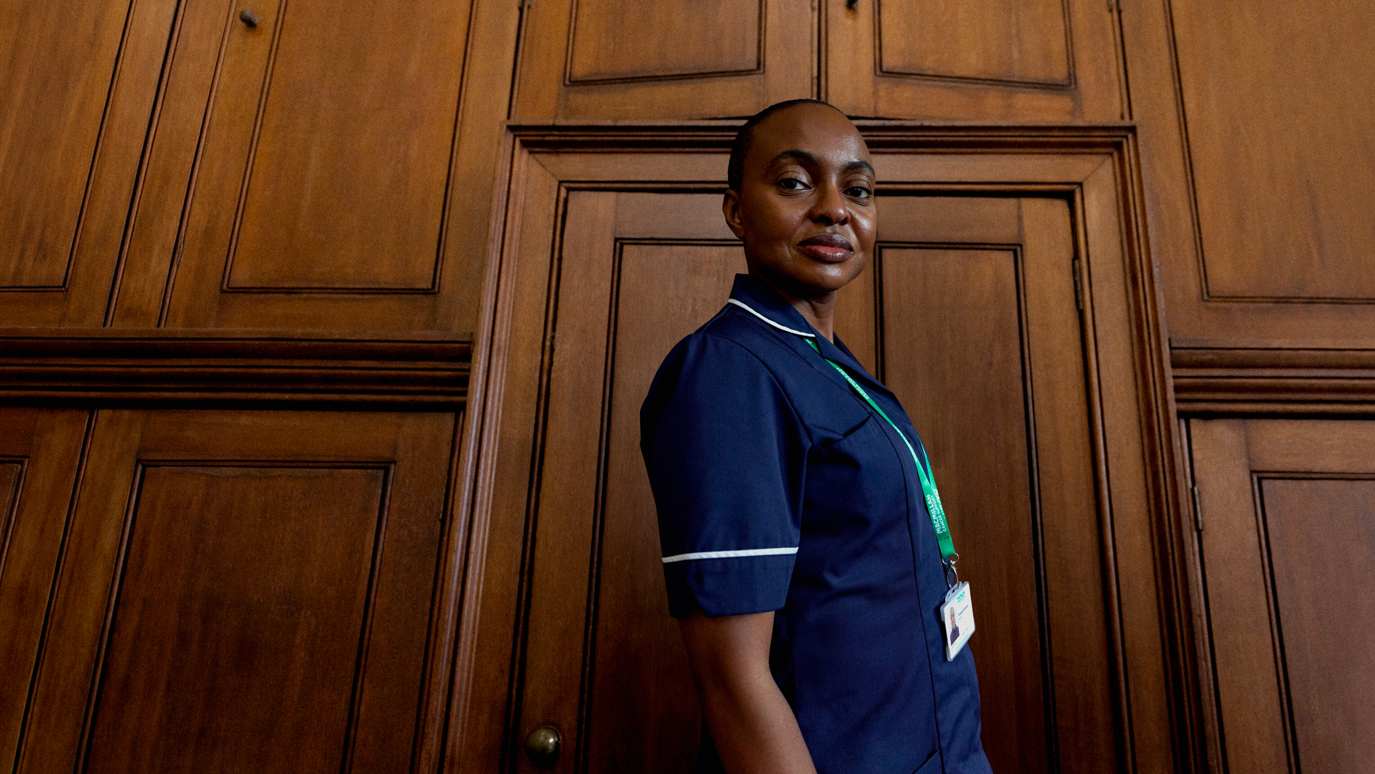 A nurse stands against a wood‑panelled wall, wearing a uniform and lanyard in a professional indoor setting.