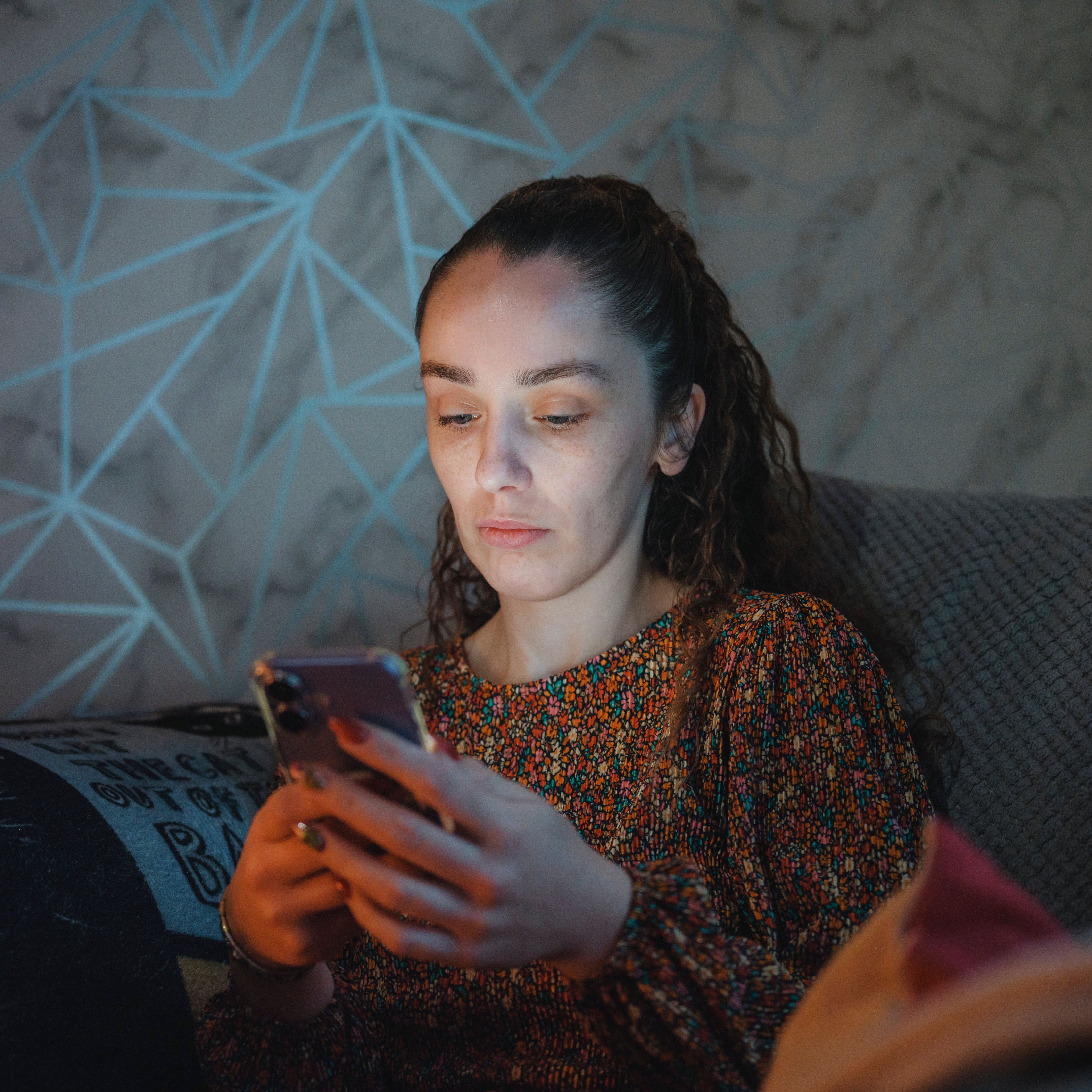 A person sits on a sofa at home, holding a smartphone and looking at the screen.