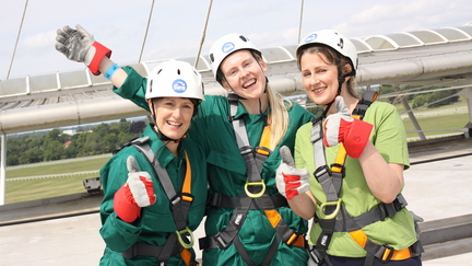 A group of three people are standing side by side after doing an abseil. They are wearing gear for the abseil including helmets, gloves and harnesses.