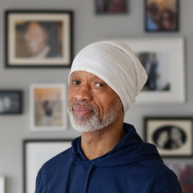 A person wearing a head covering stands indoors in front of a wall filled with framed photographs.