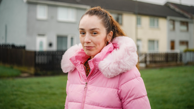 A person wearing a pink padded coat stands outdoors on a grassy area in front of residential houses.