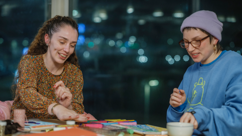 Two people sit at a table covered with colourful craft materials, working together on an art activity indoors at night.