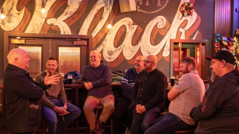 A small group of people stand together in a pub, talking under colourful string lights.