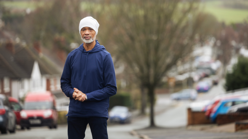 A person stands on a quiet residential street with parked cars and trees in the background.