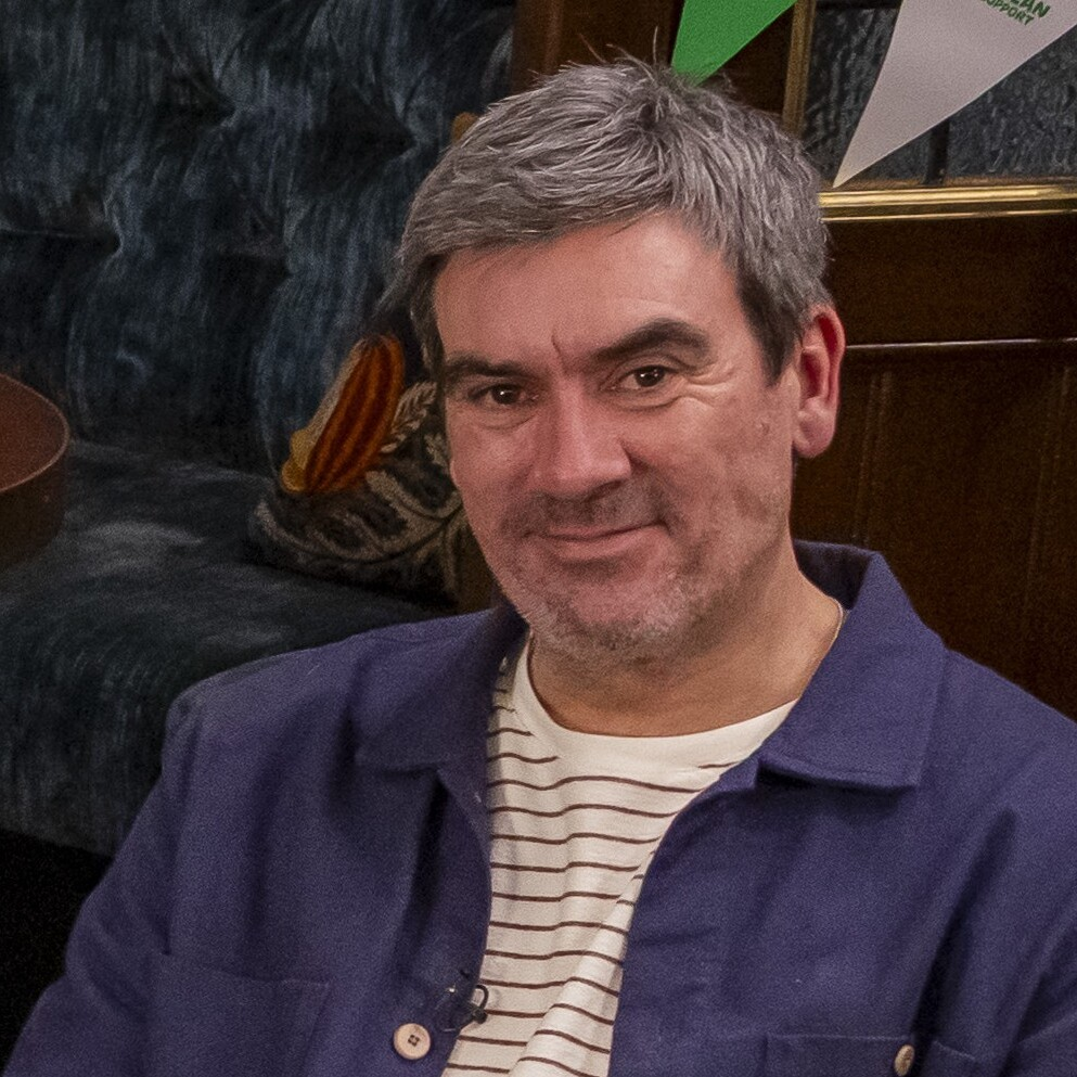 A smiling man seated in a pub-style setting, with tables, seating and Macmillan-branded bunting behind him.