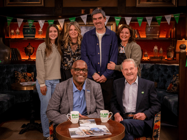 Six people in pub setting and smiling. Two are sitting at a table with the other four people standing behind them. There are Macmillan mugs and booklets on the table.