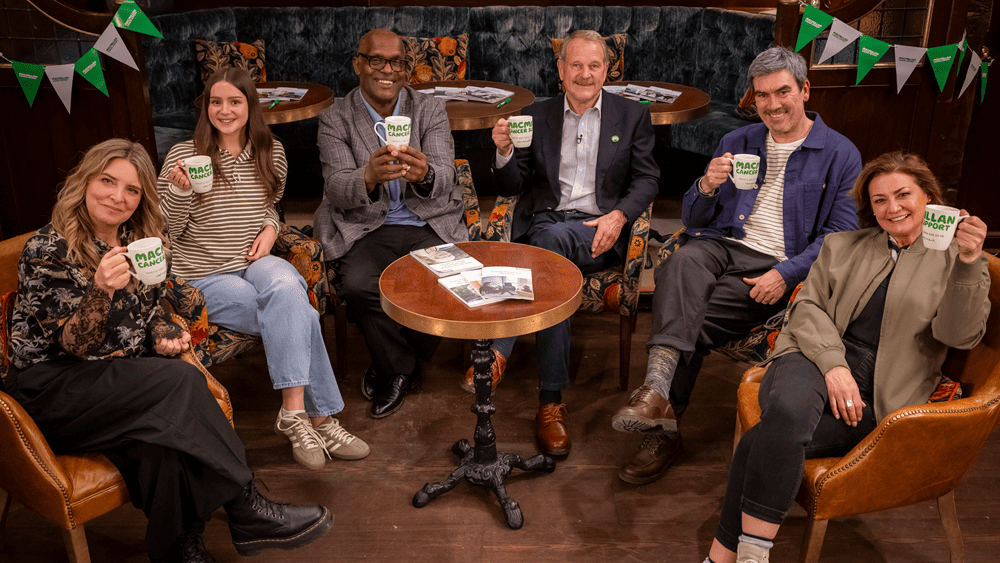 Six people sitting in a pub setting, smiling and holding Macmillan Coffee Morning mugs around a small table.