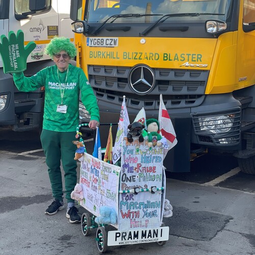 John is standing with his pram in front of a large, yellow lorry. John is wearing a bright green wig, green Macmillan long sleeved top, trousers and trainers. In one hand, John is holding up a foam glove.
