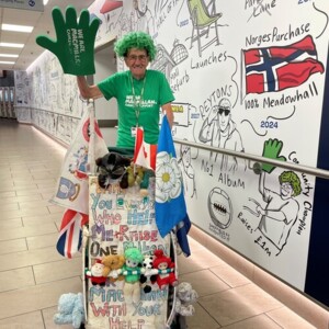John is standing behind his pram at the Meadowhall shopping centre in Sheffield. He is wearing a bright green wig and a Macmillan branded green T-shirt. In one hand he is holding up a big green foam hand. 