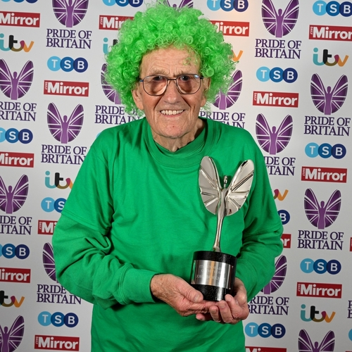 John is standing in front of a Pride of Britain backdrop. He is wearing a bright green curly wig and a green long sleeved top. In his hands he is holding a trophy. 