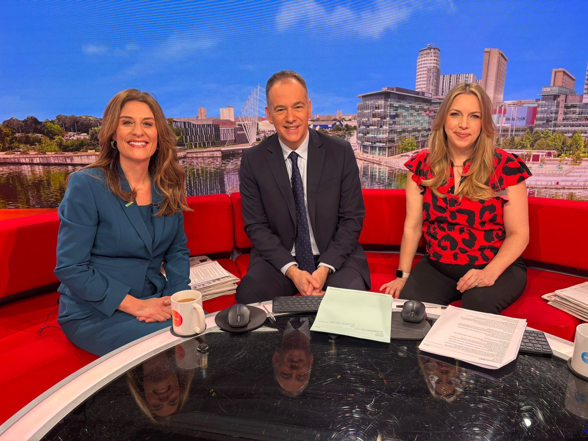 Three presenters sit on a red sofa in a television studio, with a city waterfront skyline displayed on the screen behind them.
