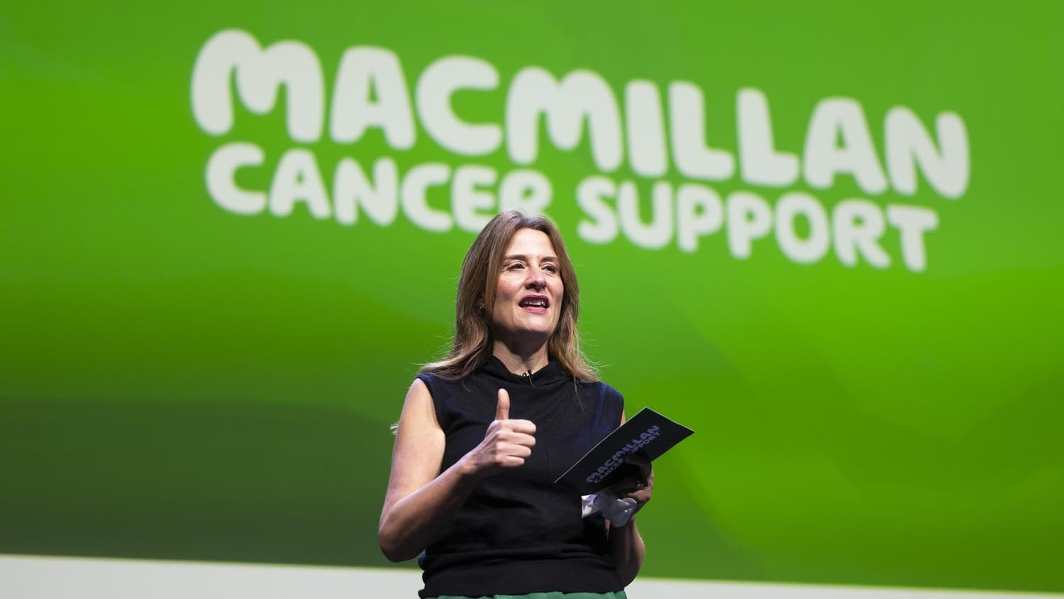 A person stands on stage holding notes and giving a thumbs‑up, with “Macmillan Cancer Support” displayed on a large green screen behind them.