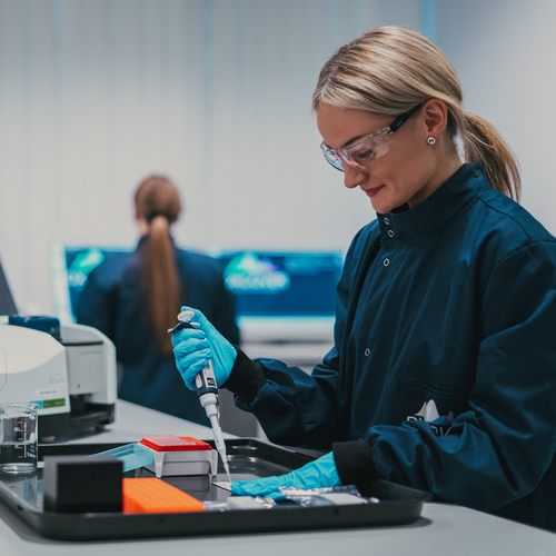 A scientist is using a transfer pipette in a laboratory. They are wearing a dark blue lab coat and safety goggles. Behind them is other lab equipment like computers.