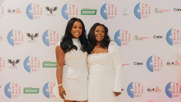 Ashleigh and Judith Neptial from From Me To You. They are standing next to each other, with one around the other. They are both wearing white clothing and standing in front of a background for an event. 