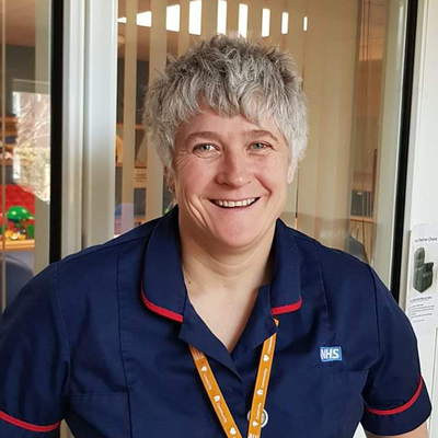 Becky is wearing a blue NHS uniform. She has short hair and is smiling. She also has on a bright orange lanyard. 