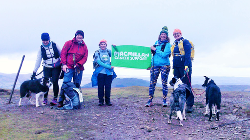 Becky is standing with her friends at completing a hike. She is holding up a green Macmillan sign with a friend. They are all wearing hiking gear and a few are partnered with their pet dogs.