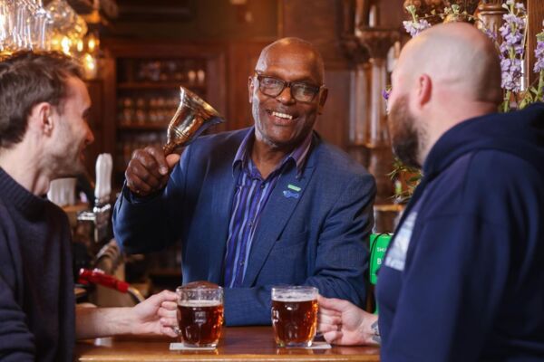 Errol, a cancer campaigner, is behind a bar in a pub ringing a bell. Two people are at the bar drinking pints of beer.