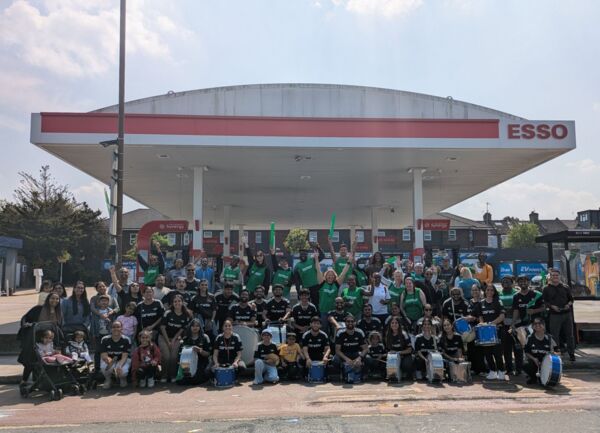 A large crowd of people in front of an ESSO garage. They have drums and banners ready to cheer on marathon runners. Some people are wearing Macmillan T Shirts, others are wearing black sports tops. 