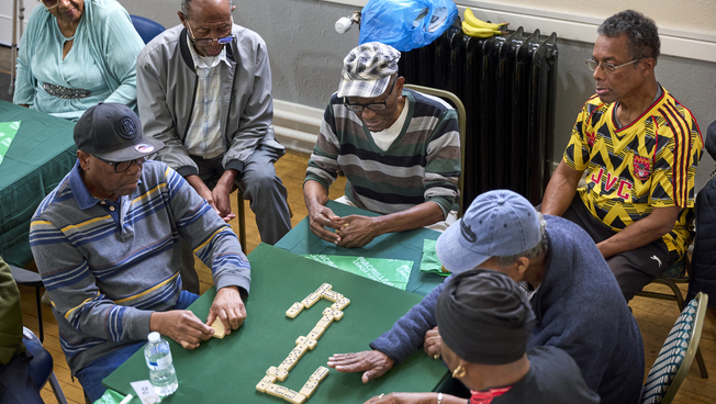 A group of men are sat around a table playing dominoes at a Coffee Morning. Some people are watching the game.