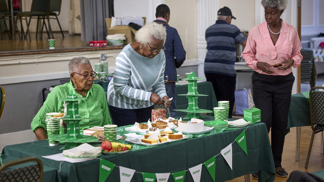 A table has been decorated for a Coffee Morning at the Pineapple Club. The table has different treats on it. Two women are behind the table and another is walking up to approach it.