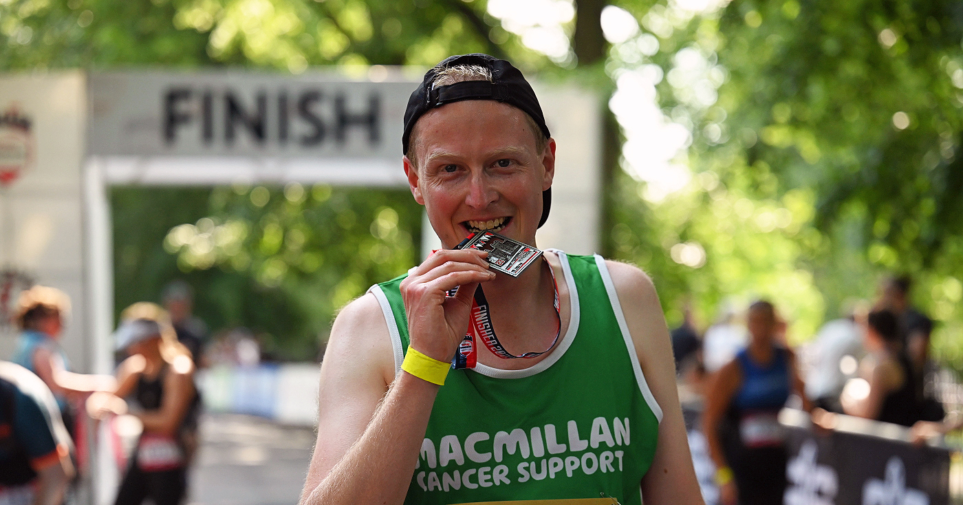 A man wearing a Macmillan vest and a cap backwards biting his finisher's medal