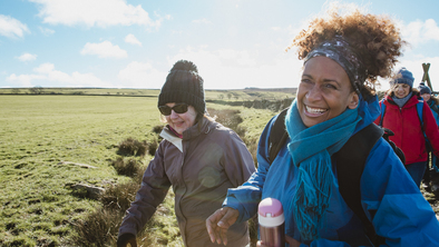 A group of women are hiking along a grassy field. They are wearing clothing for cold weather including scarves, hats and jackets. 