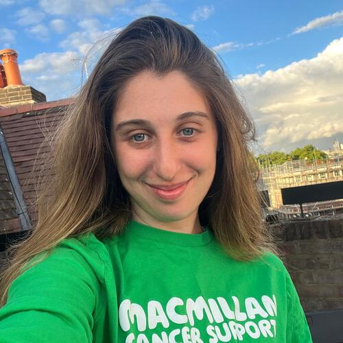 A selfie of Lola. She has long brown hair and is wearing a bright green Macmillan branded T-shirt. She is standing outside and smiling.