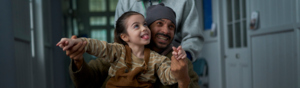 A father is sitting down in a wheelchair that is being pushing through a corridor. His daughter is in his lap and they are holding hands in the air. 