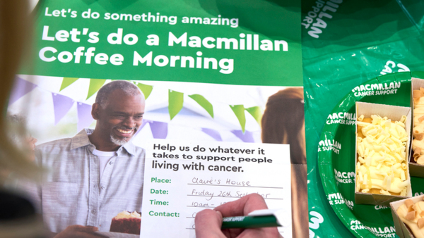 A person writing on a Coffee Morning poster with the details of their event. The poster is on a table decorated for a Coffee Morning. 