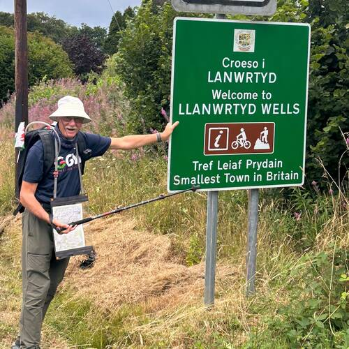 John is standing outside and learning on a green sign with information in Welsh and English stating Welcome to Llanwrtyd Wells. John is wearing a white cap, dark blue T-shirt and trousers.
