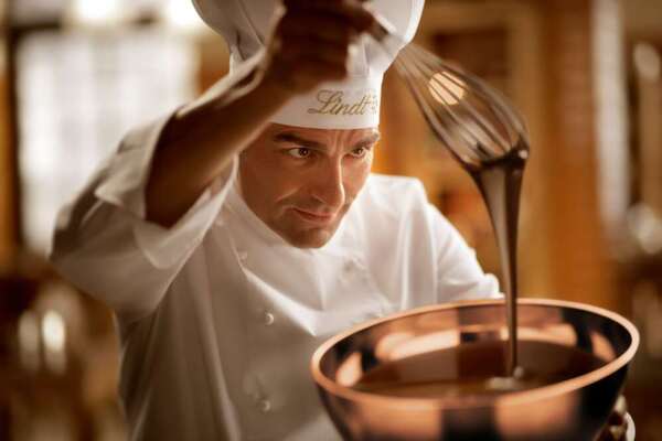 A Lindt chocolatier is holding a whisk over a bowl of melted chocolate and watching as it drips.