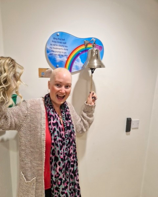 Sarah is standing next to a bell in a hospital. She is ringing it with one hand and in her other hand is a wig. Sarah is wearing a cardigan, colourful scarf and a red top.