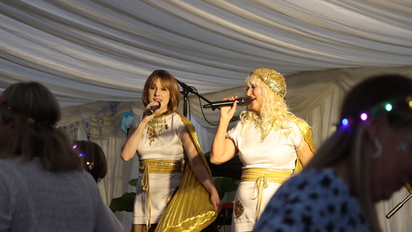 Two people are dressed up in fancy dress of Agnetha Fältskog and Anni-Frid Lyngstad of famous Swedish pop group ABBA. They are performing on a stage at a fundraising event.