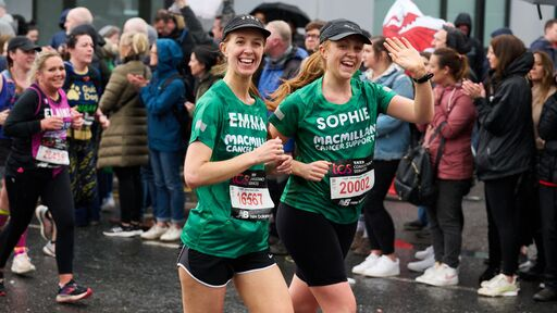 Two people are running together at an event. They are both wearing Macmillan branded green tops, caps and running shorts. People behind them are cheering on participants.