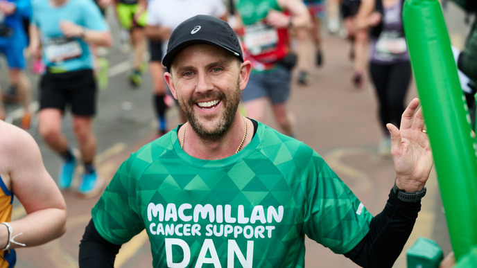 A participant taking part in a running event. They are wearing a green Macmillan top and a black cap. Behind them there are others taking part in the event.