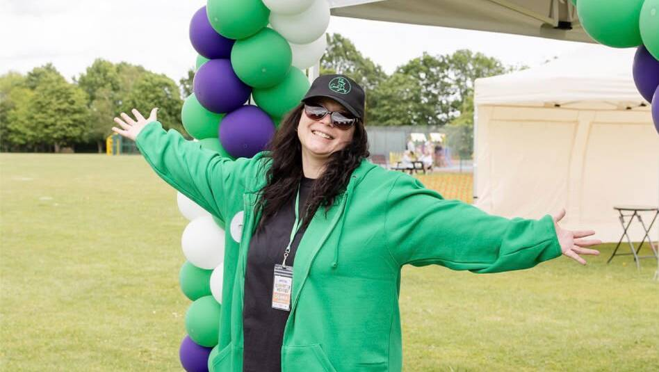 Donna is standing outside in a field of grass with her arms raised out to her sides. She is standing underneath a green, white and purple balloon arch. Donna is wearing a green hoodie, and black top, trousers and cap.