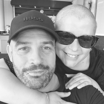 A selfie of Donna and her partner. She is behind them and has her arm around their shoulder. Their partner is wearing a dark cap and Donna is wearing sunglasses.