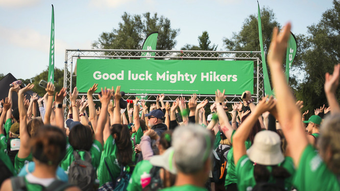 A crowd of people celebrating at a Mighty Hikes event. Their arms are in the air and in the distance is a banner that states: Good Luck Mighty Hikers.