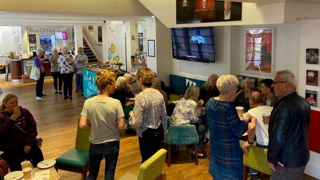 Attendees of the Oxford Playhouse's Coffee Morning. They are spread out across the venue, chatting with one another and enjoying cups of tea and coffee.