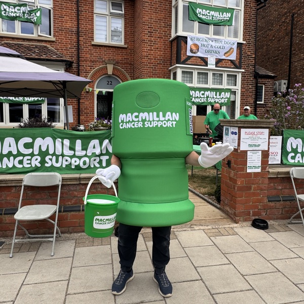 A person in a Macmillan collection tin costume. They are standing in front of Sue and John's house at the Bedford River Festival. The person in the costume is also holding a Macmillan branded collection bucket.
