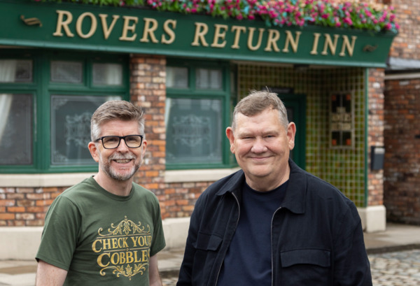 Two people outside the Rovers Return Inn on the Coronation Street set supporting Macmillan Cancer Support's Check Your Cobbles campaign.